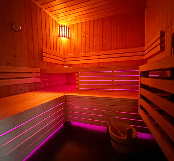 Elektrisch beheizte Sauna der Saint-Hubertus-Suite. Auf der rechten Seite befindet sich der Ofen, der mit einem Holzgeländer geschützt ist. Geradeaus und auf der linken Seite des Raumes eine geräumige hölzerne Saunabank, die auf einer Seite aus zwei Etagen besteht. Oben in der Ecke steht eine Saunalampe. Auf dem Boden steht ein Eimer mit Aufgusswasser. Unter den Saunabänken befindet sich eine farbige Beleuchtung.